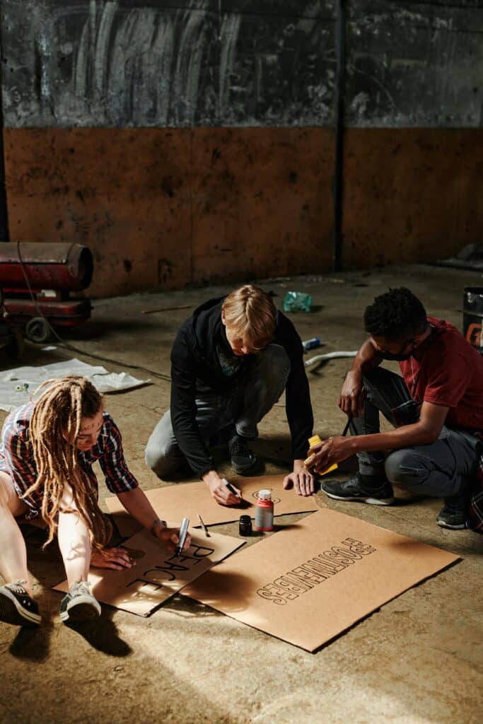 Three people creating protest signs indoors with cardboard and markers.